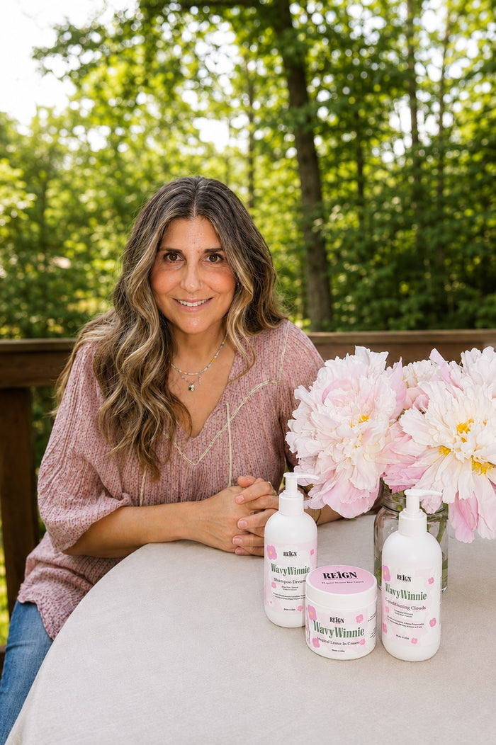 Stephanie Allyn Pohl, founder of REIGN, smiling outdoors with REIGN's The Full Crown Set curl care collection and pink peonies